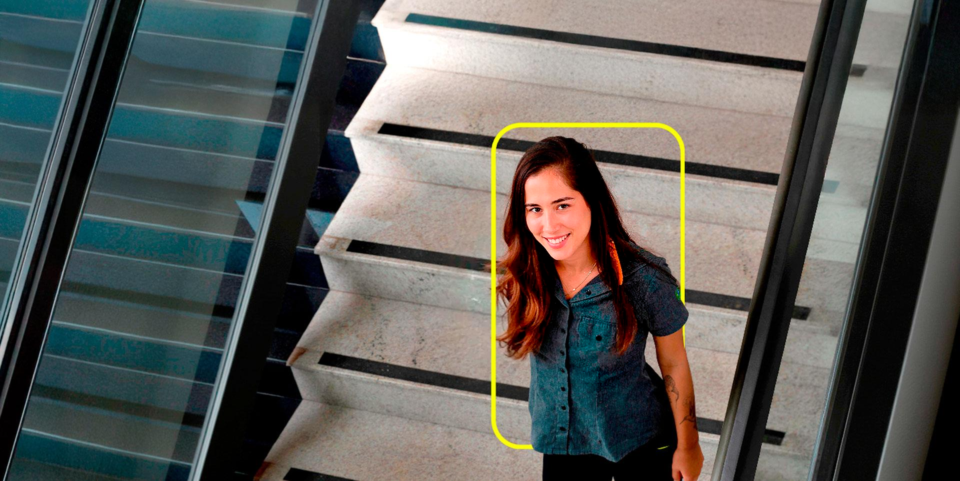 Uma jovem sorridente com cabelos longos e castanhos está olhando para a câmera. Ela veste uma camiseta escura e está parcialmente emoldurada por um contorno amarelo claro que sugere uma interface digital. Ao fundo, desfocado, há uma escada com iluminação suave.