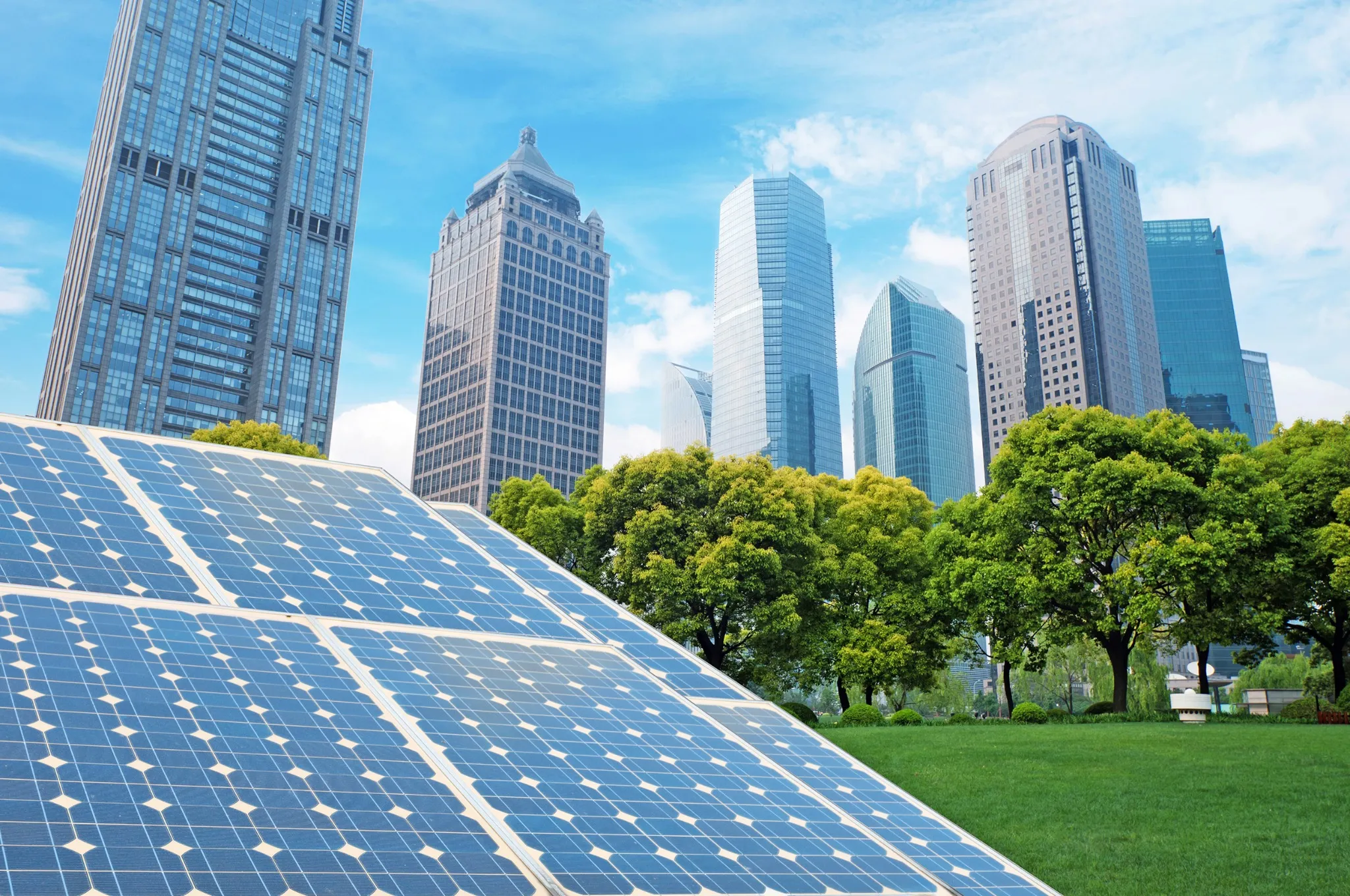 Imagem de painéis solares azuis instalados em um telhado, com reflexos do céu azul e algumas nuvens. Ao fundo, arranha-céus modernos de vidro e aço se elevam acima de árvores verdes exuberantes.