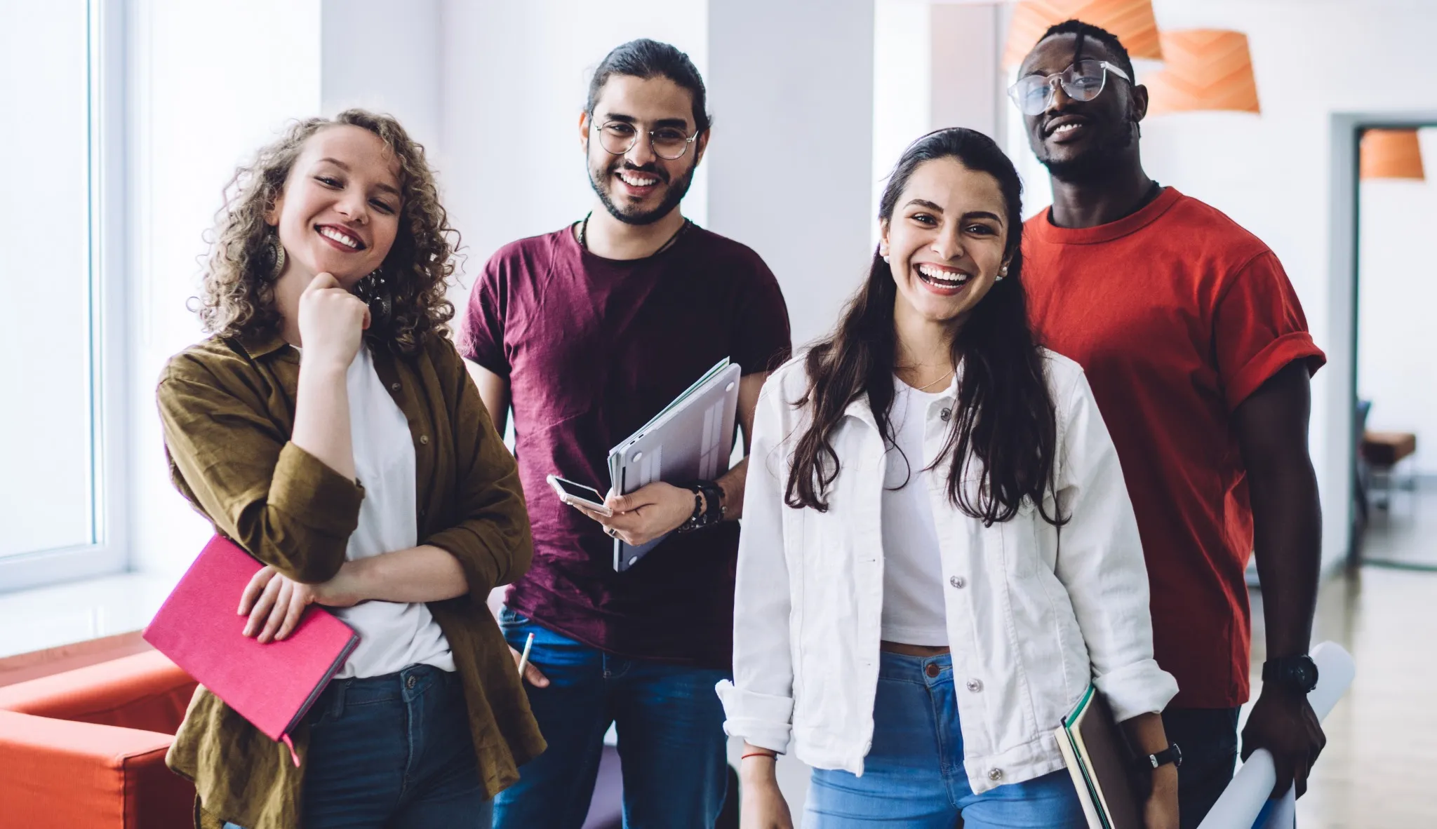 Uma equipe de estudantes de diferentes etnias, com cadernos nas mãos  e sorrindo para foto.