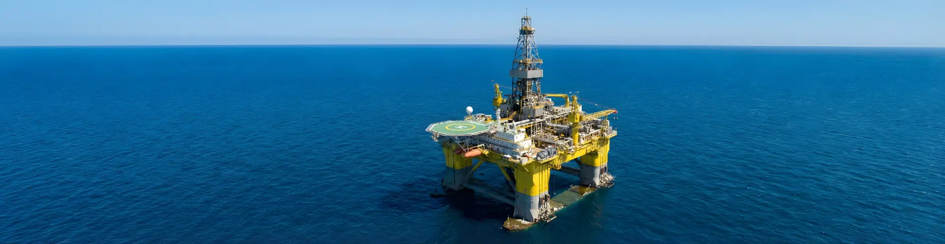 Plataforma de petróleo em operação no meio do oceano, com estrutura amarela e heliponto sobre o mar azul em dia de céu claro.