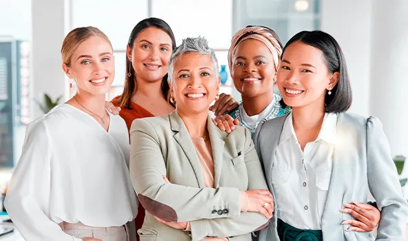 Retrato de cinco mulheres executivas de diferentes etnias sorrindo para a foto, sendo uma branca, uma latina, uma parda, uma negra e uma asiática.