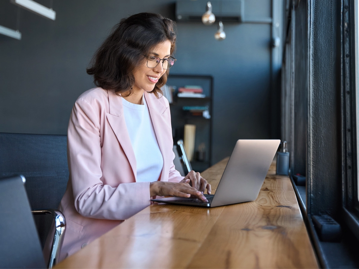 Mulher de blazer rosa digitando em notebook, representando busca por fornecedores ou oportunidades no setor de hidrogênio.